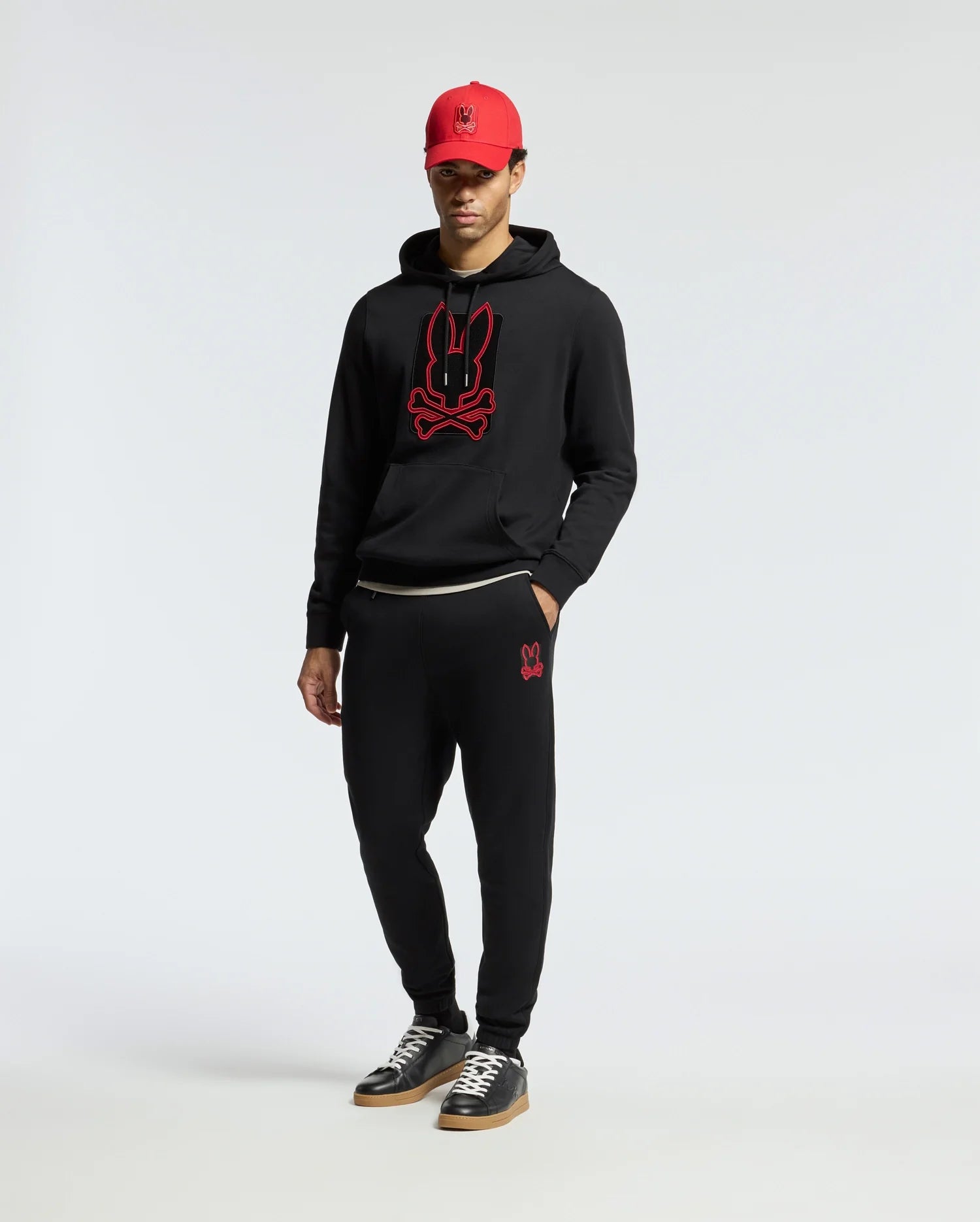 A person wearing a black hoodie with a red bunny logo, Psycho Bunny's MENS PIERCE ESSENTIAL FRENCH TERRY SWEATPANT - B6P136E200 featuring a small bunny design, and a red cap. They are standing against a plain white background and wearing black sneakers.