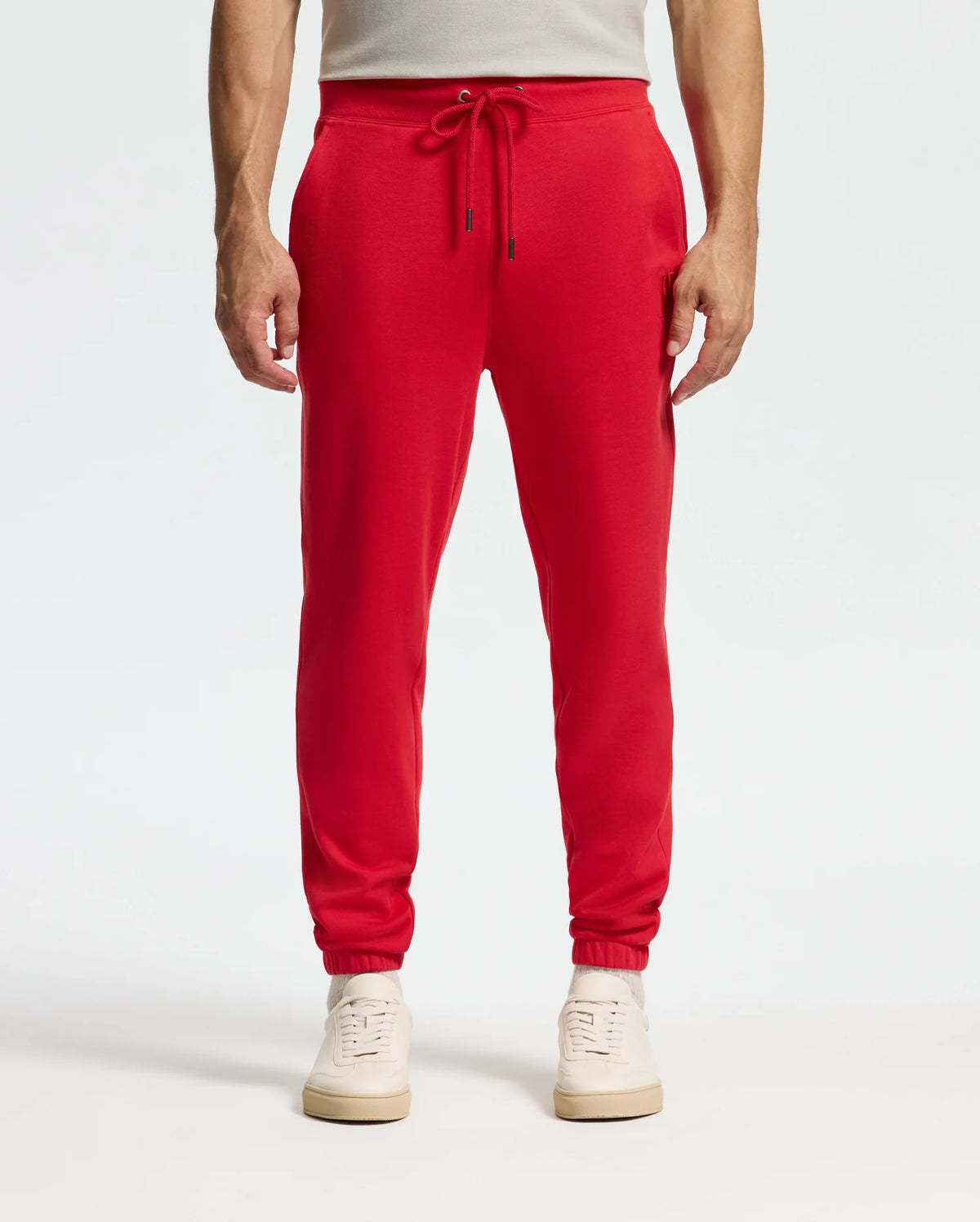 A person is dressed in Psycho Bunny's vibrant red *Mens Pierce Essential French Terry Sweatpant - B6P136E200*, combined with white sneakers. These joggers, designed with a drawstring waist and pockets, are highlighted against a simple, light-colored backdrop, showing just the lower half of their body.