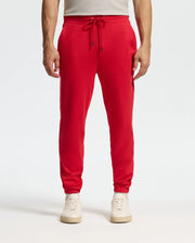 A person is dressed in Psycho Bunny's vibrant red *Mens Pierce Essential French Terry Sweatpant - B6P136E200*, combined with white sneakers. These joggers, designed with a drawstring waist and pockets, are highlighted against a simple, light-colored backdrop, showing just the lower half of their body.