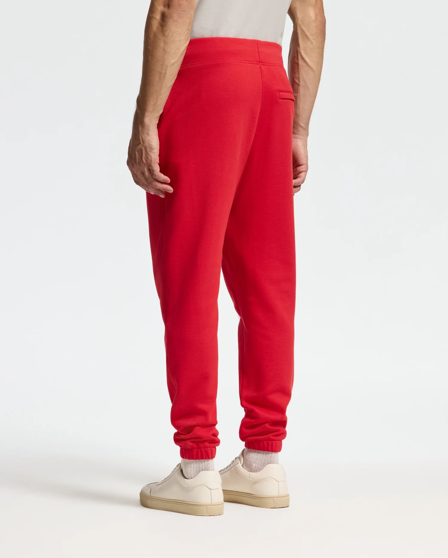 A person dressed in the Psycho Bunny MENS PIERCE ESSENTIAL FRENCH TERRY SWEATPANT - B6P136E200 in vibrant red pairs it with a light gray top, and completes the look with white sneakers and socks, all set against a plain, bright background.
