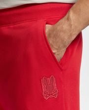 A close-up shows someone wearing the Psycho Bunny MENS PIERCE ESSENTIAL FRENCH TERRY SWEATPANT - B6P136E200 in vibrant red, casually tucking their hand into the pocket. These cotton French terry joggers boast an embroidered patch featuring a bunny with crossed bones. The neutral background highlights their bold style.