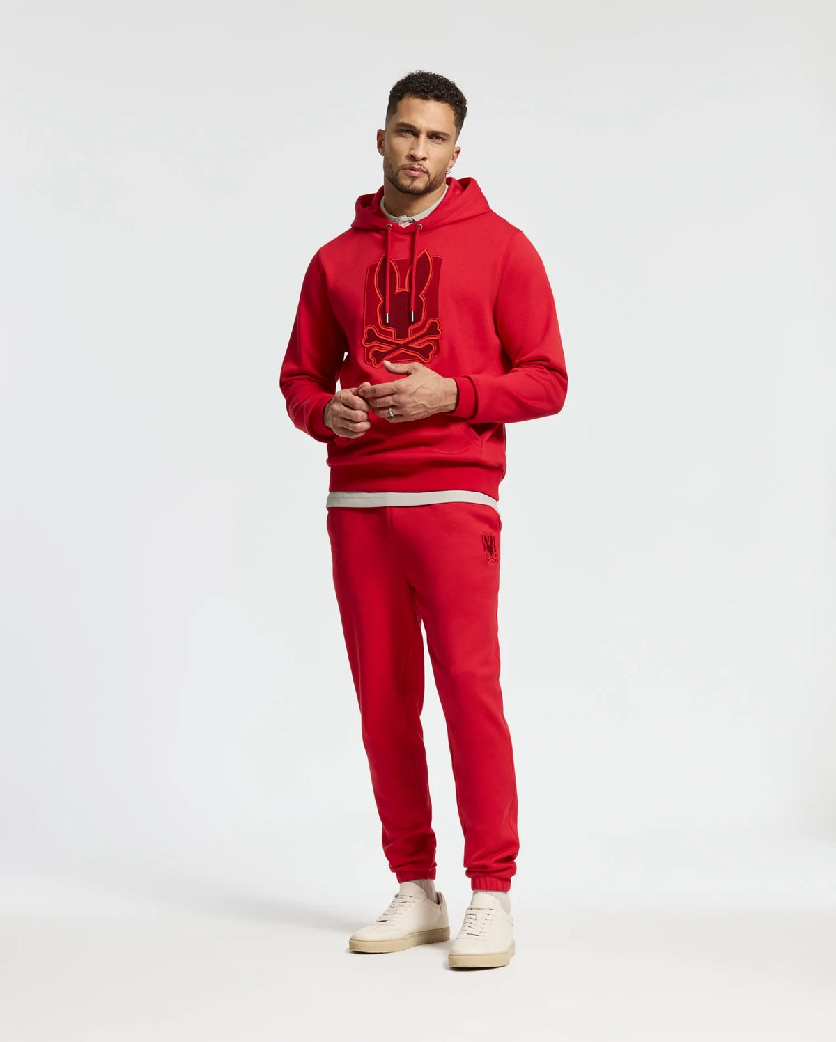 A man stands wearing a red hoodie with a graphic design, paired with matching Psycho Bunny MENS PIERCE ESSENTIAL FRENCH TERRY SWEATPANTS (B6P136E200), and white sneakers against a plain background.