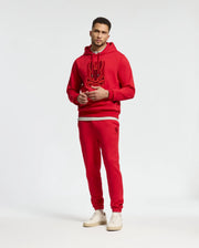 A man stands wearing a red hoodie with a graphic design, paired with matching Psycho Bunny MENS PIERCE ESSENTIAL FRENCH TERRY SWEATPANTS (B6P136E200), and white sneakers against a plain background.