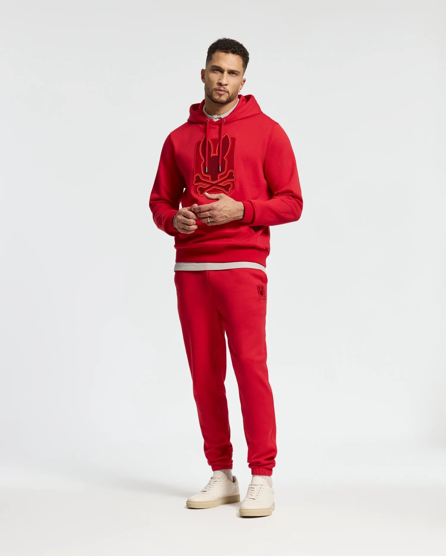 A man stands wearing a red hoodie with a graphic design, paired with matching Psycho Bunny MENS PIERCE ESSENTIAL FRENCH TERRY SWEATPANTS (B6P136E200), and white sneakers against a plain background.