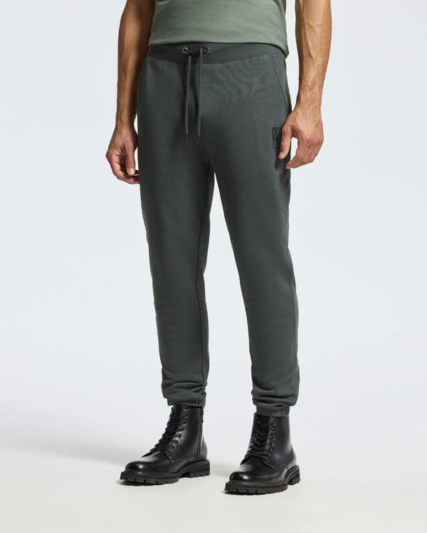 A person wearing the dark green Psycho Bunny MENS PIERCE ESSENTIAL FRENCH TERRY SWEATPANT - B6P136E200 and black combat boots stands on a neutral background. The sweatpants feature an adjustable drawstring at the waist, and the boots have a chunky sole. Only the lower half of the body is visible.