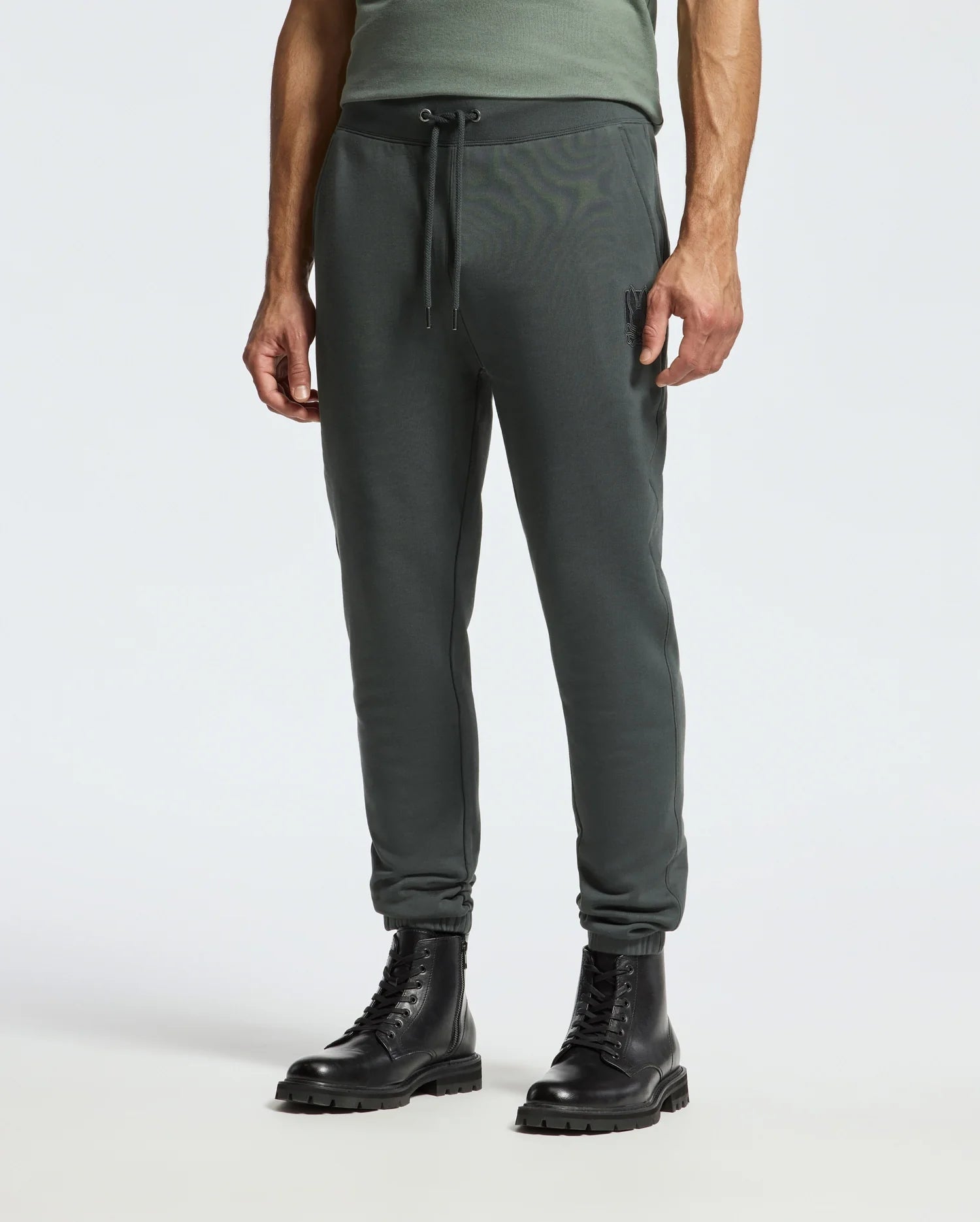 A person wearing the dark green Psycho Bunny MENS PIERCE ESSENTIAL FRENCH TERRY SWEATPANT - B6P136E200 and black combat boots stands on a neutral background. The sweatpants feature an adjustable drawstring at the waist, and the boots have a chunky sole. Only the lower half of the body is visible.