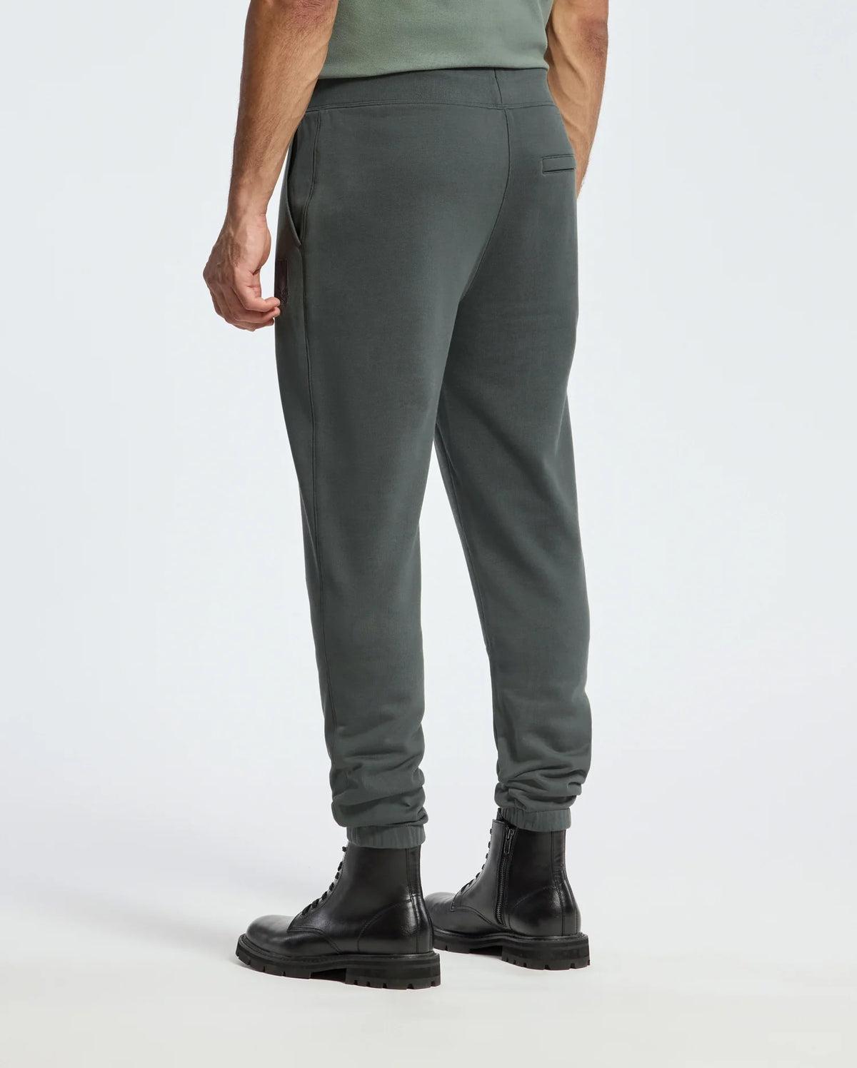 A person is wearing Psycho Bunny's Men's Pierce Essential French Terry Sweatpants in dark green, paired with black leather boots, standing against a plain white background. The sweatpants feature a slim fit with an elastic waistband and cuffs. The person is seen from the back.
