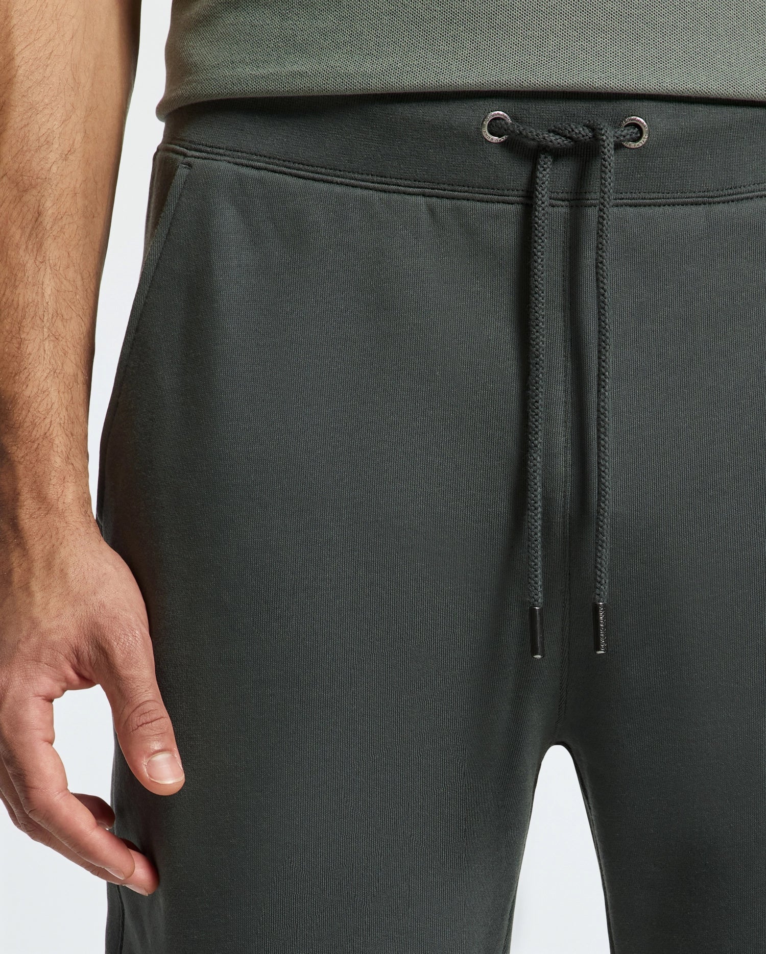 A close-up of a person wearing Psycho Bunny's MENS PIERCE ESSENTIAL FRENCH TERRY SWEATPANT in dark gray. The image focuses on the waist area, showcasing a visible drawstring, with the person's left hand resting beside the pants. The background is a simple, light gray.