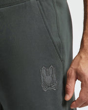 A close-up of a person's hand resting on the side pocket of dark gray sweatpants from Psycho Bunny. Crafted from cotton French terry, these Men's Pierce Essential Sweatpants feature a stitched design of a rabbit head with crossbones near the pocket.