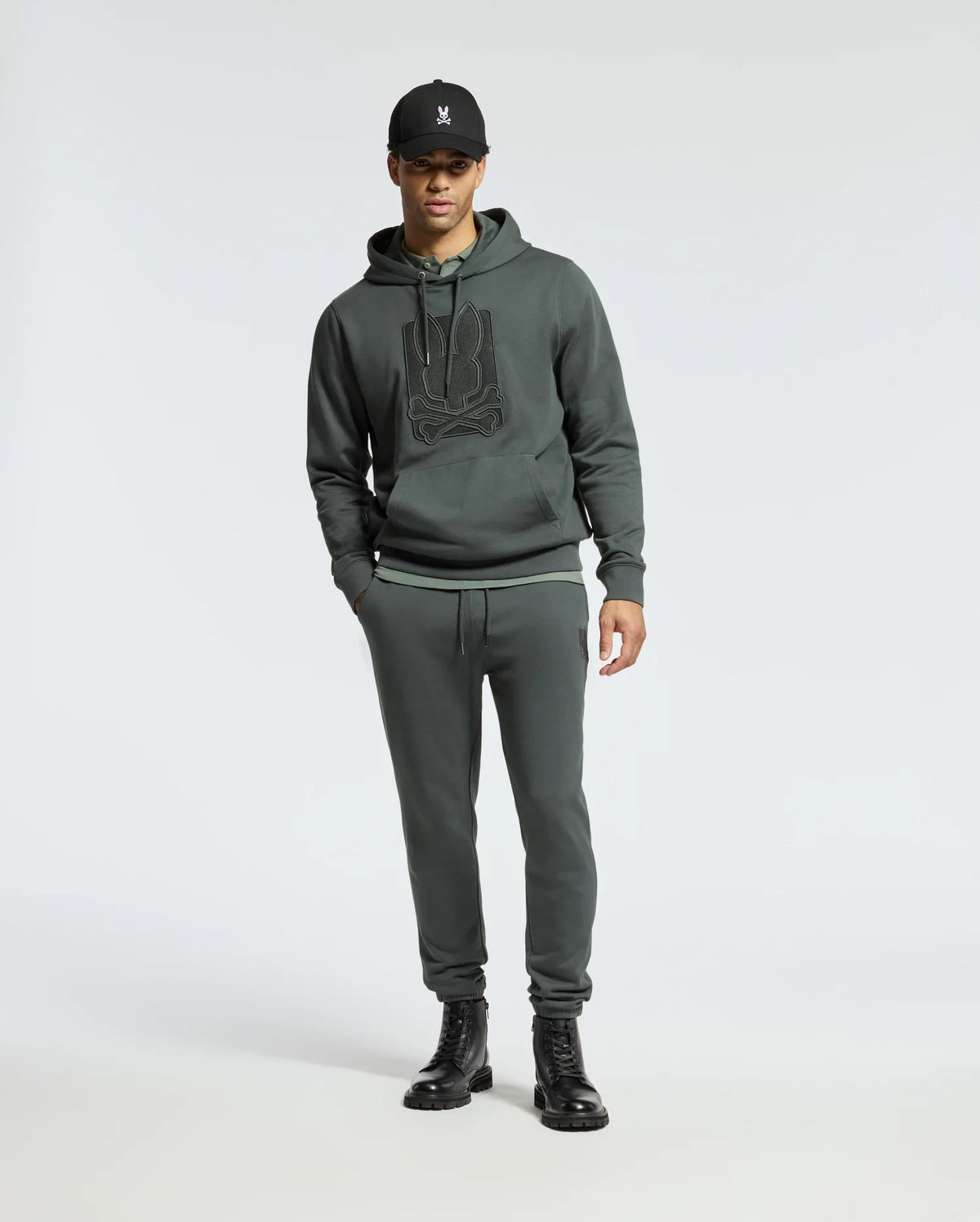 A person wearing a dark hoodie with a logo, Psycho Bunny MENS PIERCE ESSENTIAL FRENCH TERRY SWEATPANT - B6P136E200, black boots, and a black cap stands against a plain, light background.