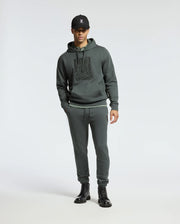 A person wearing a dark hoodie with a logo, Psycho Bunny MENS PIERCE ESSENTIAL FRENCH TERRY SWEATPANT - B6P136E200, black boots, and a black cap stands against a plain, light background.