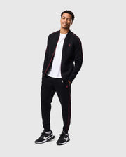 A man is standing and smiling while dressed in a black Psycho Bunny MENS TRAVIS TRACK PANT - B6P143D200 with red accents and a white T-shirt, made of high-quality cotton-blend for regular fit. He is wearing black and white sneakers. The background is plain and light-colored.