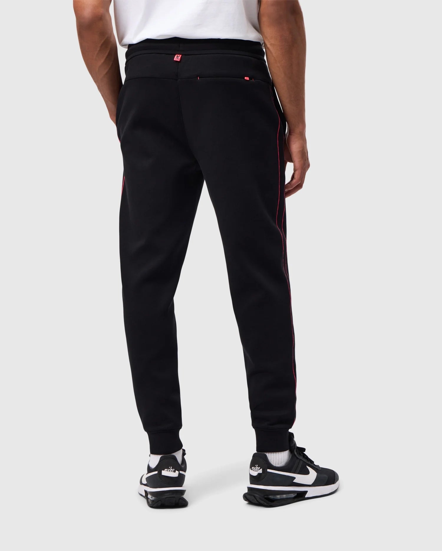 Rear view of a person wearing a high-quality cotton-blend white shirt, black athletic pants with red side stripes called MENS TRAVIS TRACK PANT - B6P143D200 by Psycho Bunny, and black and white Nike Air sneakers. The background is plain off-white.