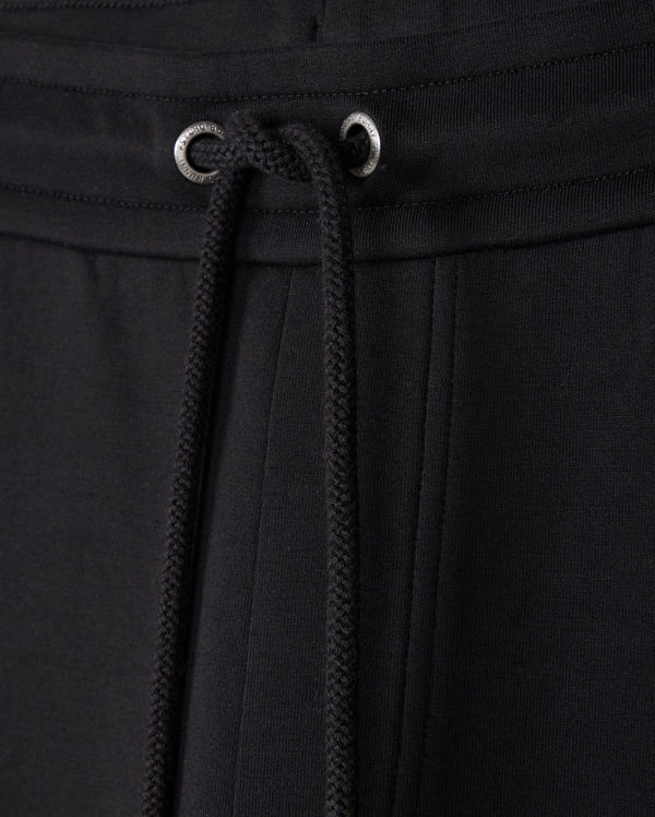 Close-up of black Psycho Bunny MENS TRAVIS TRACK PANT - B6P143D200 featuring a drawstring waistband. The drawstrings are threaded through metal eyelets, and the high-quality cotton-blend fabric has a smooth, slightly textured appearance. The stitching and seams are visible, showcasing the garment's craftsmanship.