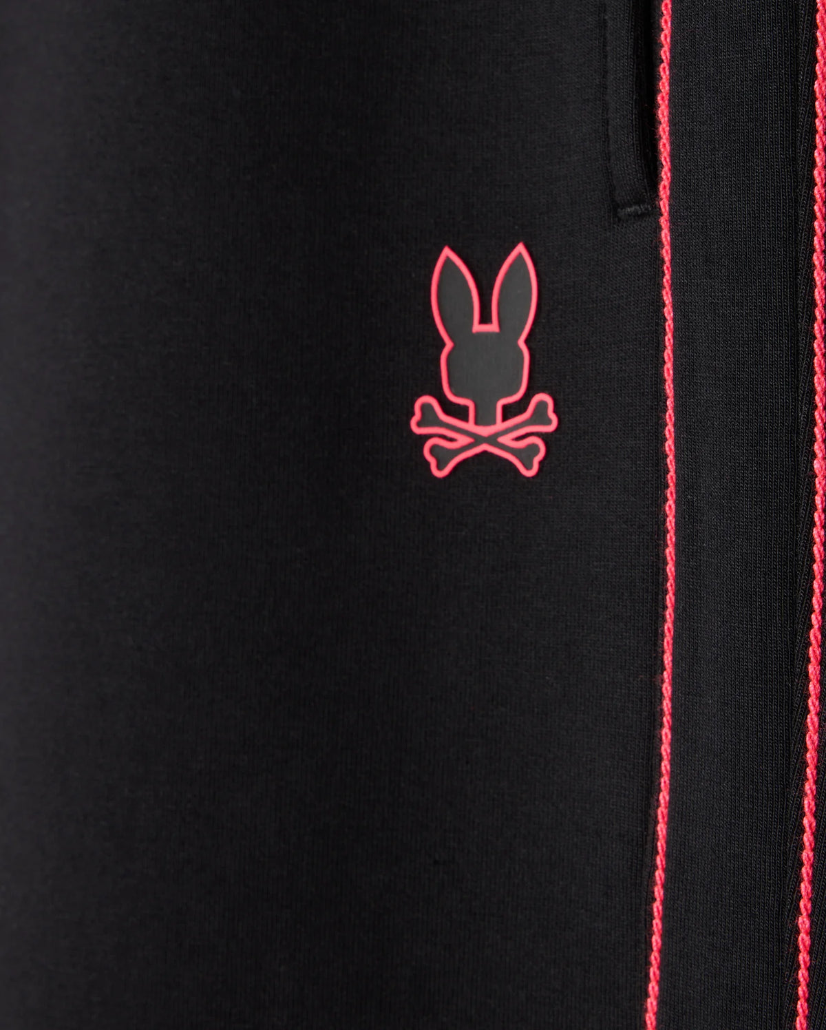 Close-up of the Psycho Bunny MENS TRAVIS TRACK PANT (B6P143D200) in black fabric, featuring red stitching detail and an embroidered pink logo of a stylized bunny head above crossbones, crafted from high-quality cotton-blend material.