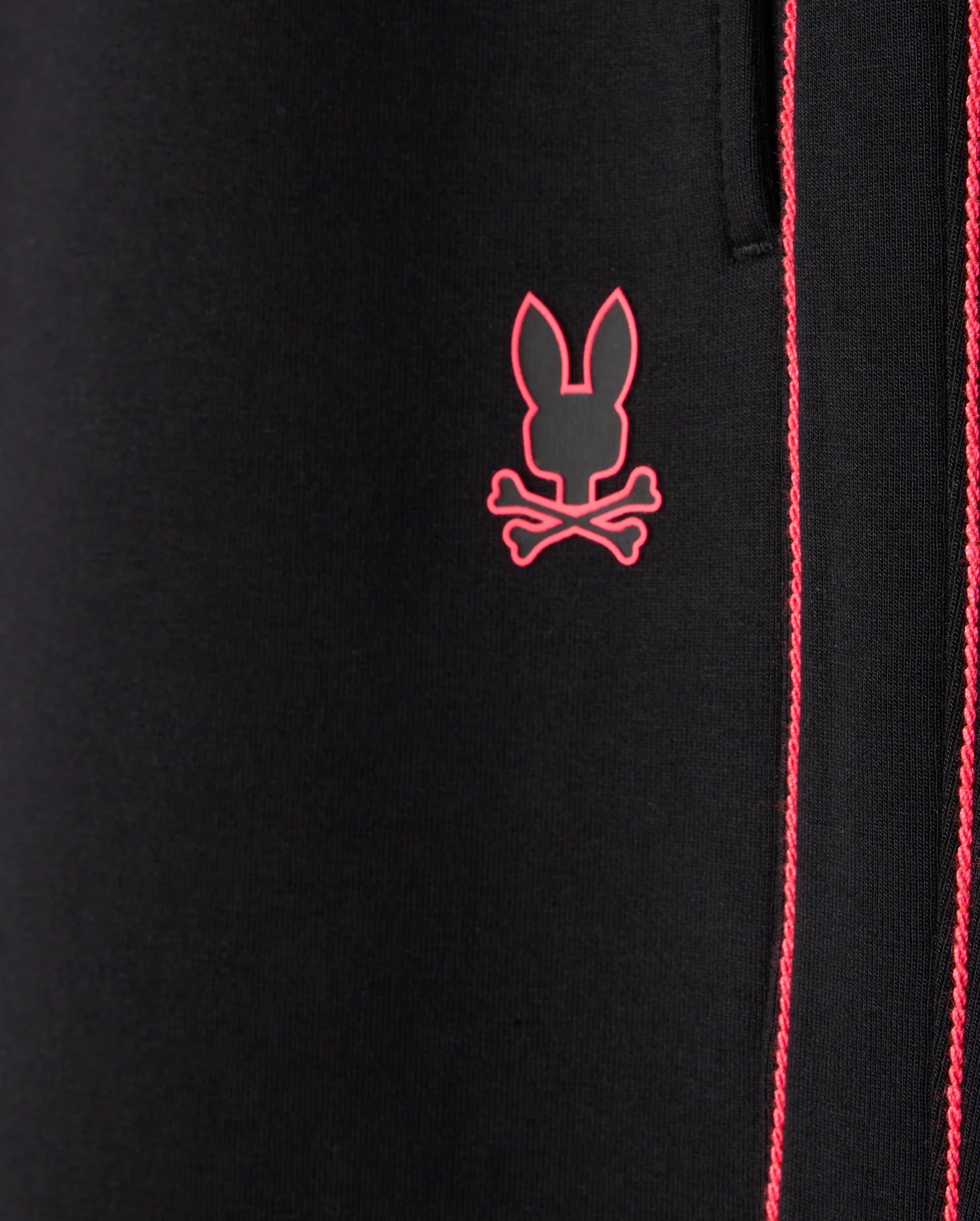 Close-up of the Psycho Bunny MENS TRAVIS TRACK PANT (B6P143D200) in black fabric, featuring red stitching detail and an embroidered pink logo of a stylized bunny head above crossbones, crafted from high-quality cotton-blend material.
