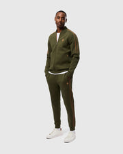 A man wearing a Psycho Bunny olive green tracksuit with orange accents, made from a high-quality cotton-blend, stands against a plain white background. He has his hands in the invisible zip pockets of the MENS TRAVIS TRACK PANT - B6P143D200 and is wearing white sneakers. The tracksuit jacket features a small embroidered logo on the left side.