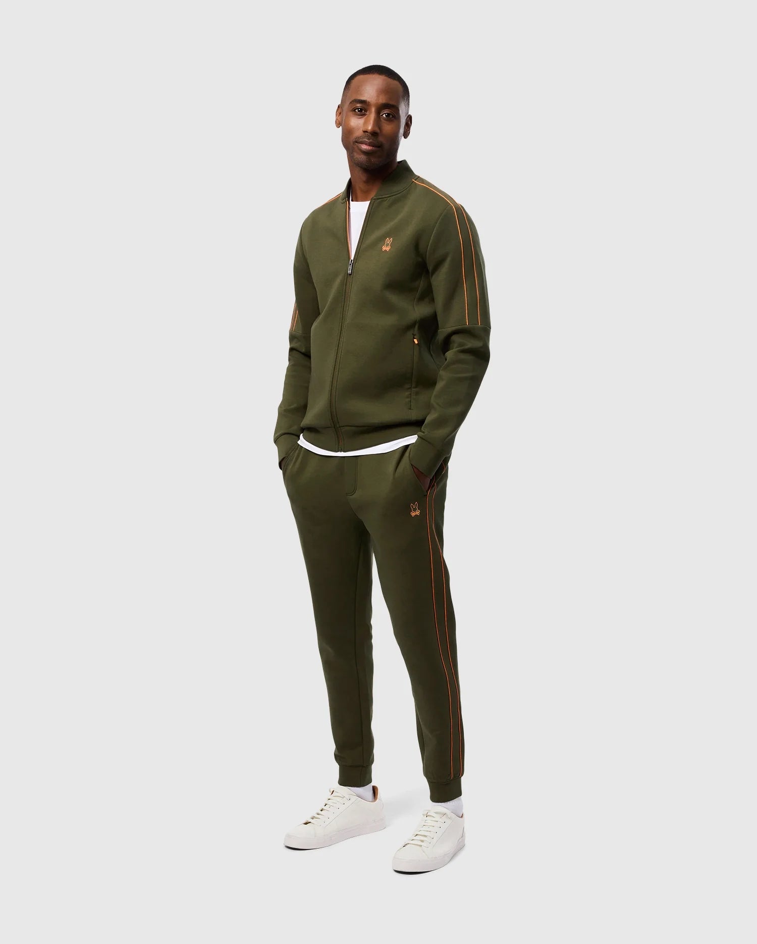 A man wearing a Psycho Bunny olive green tracksuit with orange accents, made from a high-quality cotton-blend, stands against a plain white background. He has his hands in the invisible zip pockets of the MENS TRAVIS TRACK PANT - B6P143D200 and is wearing white sneakers. The tracksuit jacket features a small embroidered logo on the left side.
