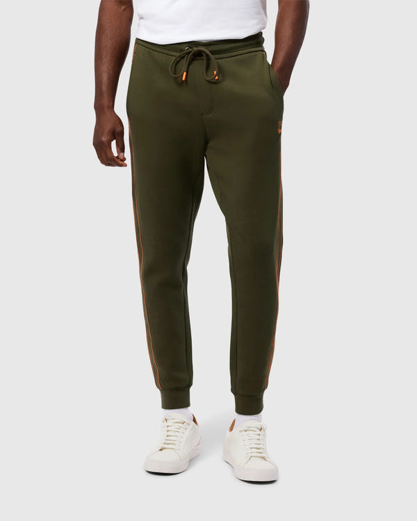 A person is wearing Psycho Bunny's MENS TRAVIS TRACK PANT - B6P143D200 in olive green with orange side stripes, a white t-shirt, and white sneakers with brown accents. The high-quality cotton-blend jogger pants feature a drawstring waist and invisible zip pockets. The person's hands are relaxed at their sides, standing against a plain background.