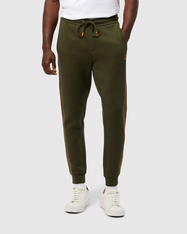 A person is wearing Psycho Bunny's MENS TRAVIS TRACK PANT - B6P143D200 in olive green with orange side stripes, a white t-shirt, and white sneakers with brown accents. The high-quality cotton-blend jogger pants feature a drawstring waist and invisible zip pockets. The person's hands are relaxed at their sides, standing against a plain background.