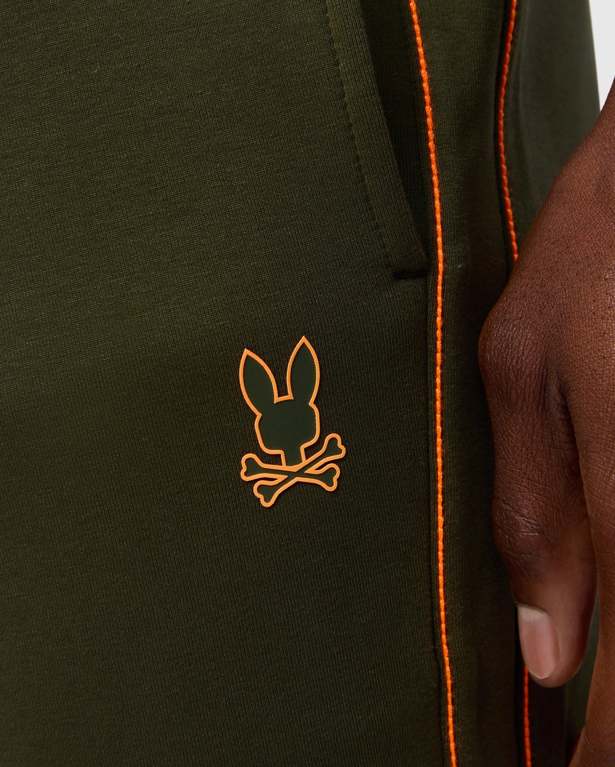 Close-up of a person wearing MENS TRAVIS TRACK PANT - B6P143D200 by Psycho Bunny, in green with orange piping along the seams. The high-quality cotton-blend pants feature invisible zip pockets and a logo of a stylized bunny head and crossbones in orange, located near the pocket. The person's hand rests beside the pocket.
