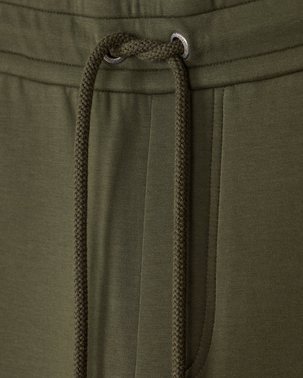 Close-up of the waistband and drawstring of a pair of olive green MENS TRAVIS TRACK PANT - B6P143D200 by Psycho Bunny. The image highlights the texture of the high-quality cotton-blend fabric and the metal eyelets through which the drawstring is threaded. A pocket is partially visible on the right side.