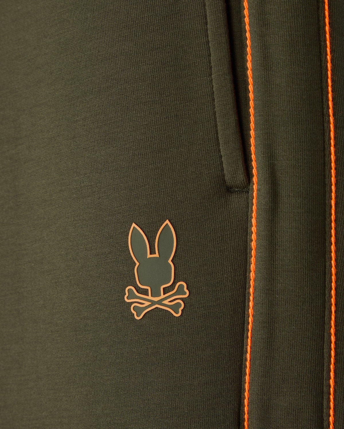 Close-up of the Psycho Bunny Men's Travis Track Pant (B6P143D200) in dark green, crafted from a high-quality cotton-blend material featuring a pocket with orange stitching. Below the pocket, there's an orange logo of a rabbit head overlaid on two crossed bones.