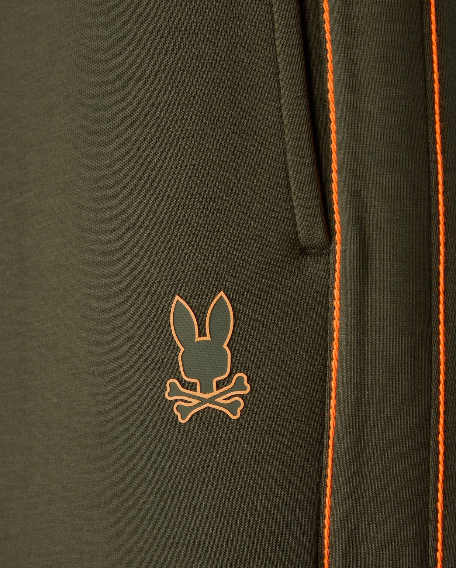 Close-up of the Psycho Bunny Men's Travis Track Pant (B6P143D200) in dark green, crafted from a high-quality cotton-blend material featuring a pocket with orange stitching. Below the pocket, there's an orange logo of a rabbit head overlaid on two crossed bones.