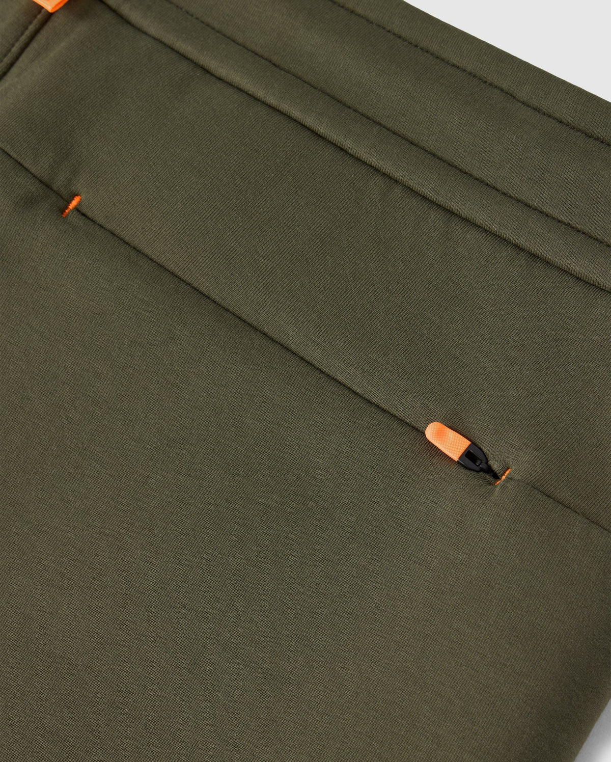Close-up of an olive green fabric, typical of the Psycho Bunny MENS TRAVIS TRACK PANT - B6P143D200, featuring a hidden zipper pocket. The orange zipper pull is partially visible, blending seamlessly with the high-quality cotton-blend material. The fabric has a smooth texture and neat stitching along the edges.