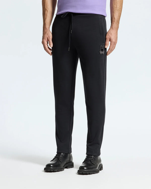 A person is wearing the MENS ALEXANDER SWEATPANT by Psycho Bunny, which features a small embroidered logo on the left thigh. These cotton joggers include an elastic waistband with a drawstring for comfort. The ensemble is completed with black shoes and a purple shirt.