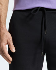 The individual is dressed in a purple shirt and the Psycho Bunny MENS ALEXANDER SWEATPANT - B6P155E200 in black, with special attention to their hand resting near the waist area of these cotton sweatpants.