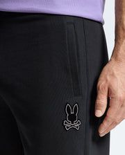 A close-up shot showcases a person wearing the MENS ALEXANDER SWEATPANT - B6P155E200 by Psycho Bunny, in black cotton. The joggers feature the distinct rabbit skull and crossbones logo near the knee. Paired with a light purple top, they stand casually with their right hand by their side, capturing the relaxed style of these comfortable men's joggers.