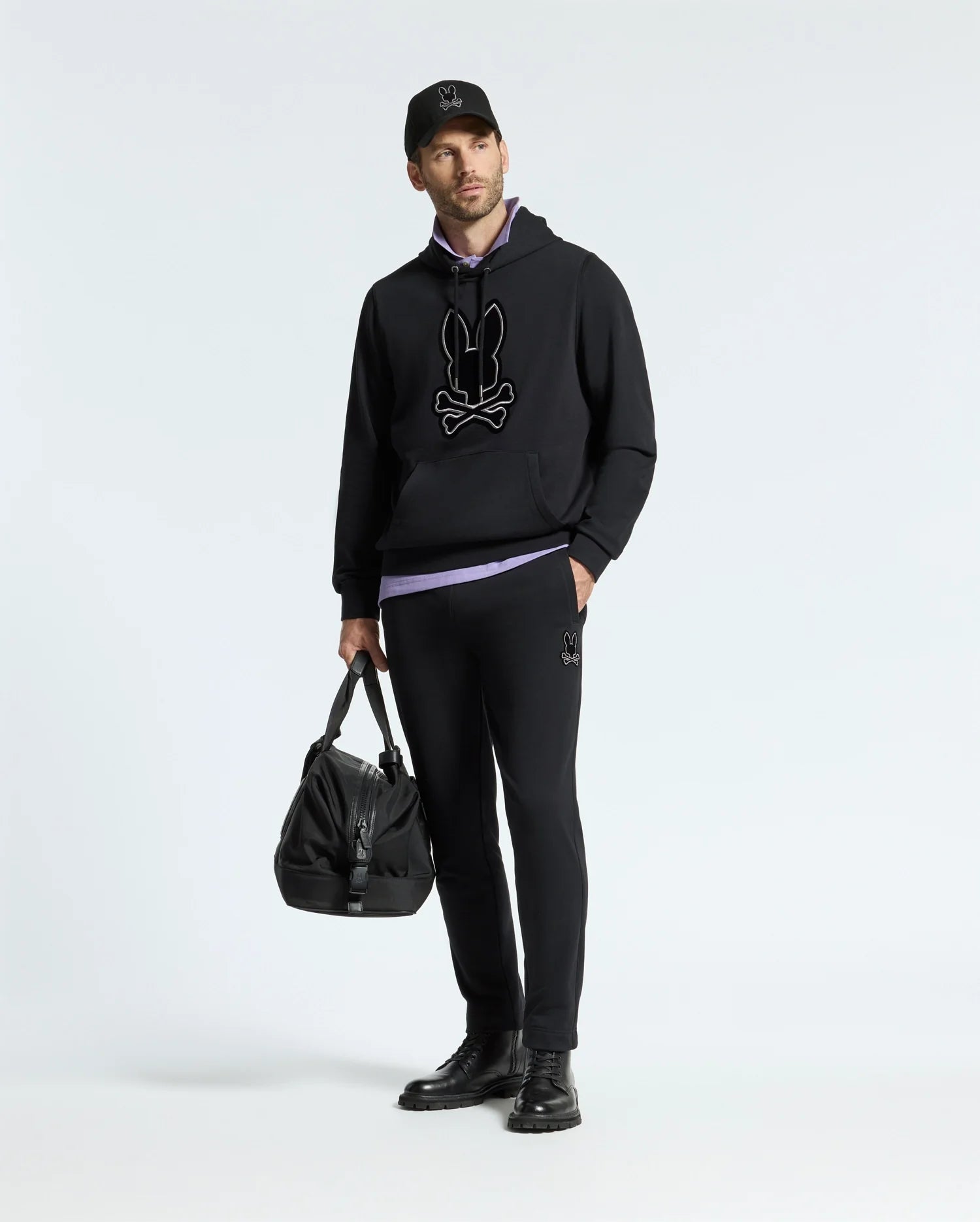 A man stands against a simple backdrop, dressed in a black hoodie featuring a graphic design, the MENS ALEXANDER SWEATPANT by Psycho Bunny, and a black cap. He is holding a large black bag and wearing black shoes.