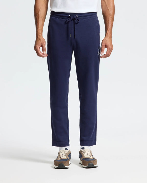 A man in Psycho Bunny's MENS ALEXANDER SWEATPANT - B6P155E200, relaxed fit navy joggers with a drawstring waist and front embroidery, paired with a white T-shirt and brown sneakers.