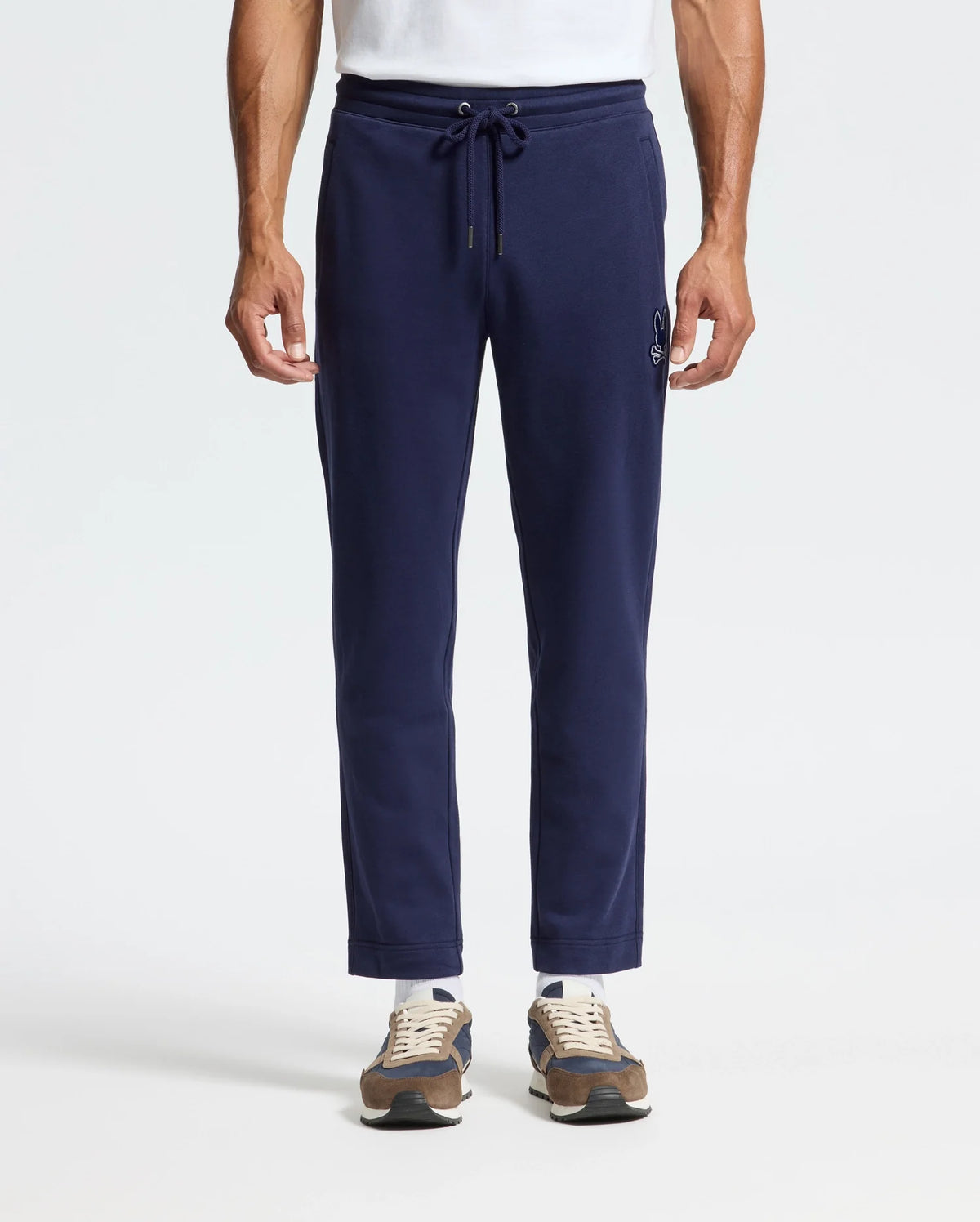 A man in Psycho Bunny's MENS ALEXANDER SWEATPANT - B6P155E200, relaxed fit navy joggers with a drawstring waist and front embroidery, paired with a white T-shirt and brown sneakers.