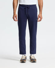 A man in Psycho Bunny's MENS ALEXANDER SWEATPANT - B6P155E200, relaxed fit navy joggers with a drawstring waist and front embroidery, paired with a white T-shirt and brown sneakers.