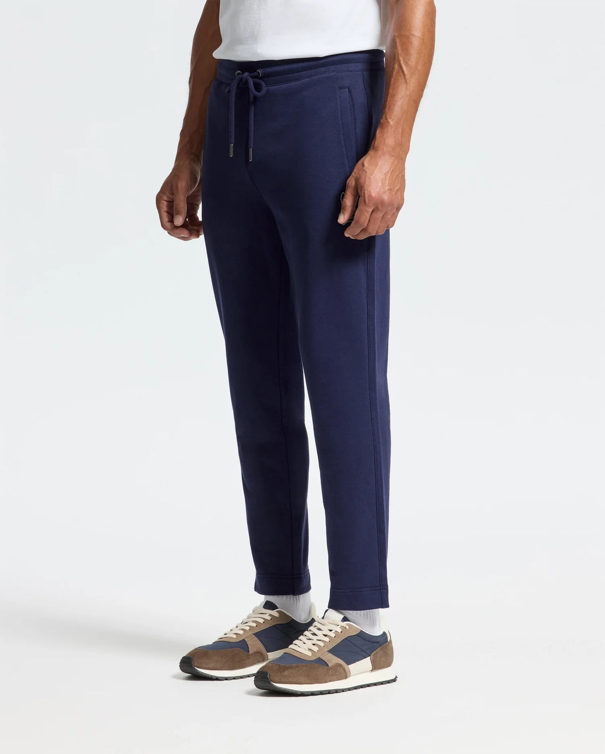 A person dressed in Psycho Bunny's MENS ALEXANDER SWEATPANT - B6P155E200, a navy cotton relaxed jogger, paired with a white shirt, blue and brown sneakers, and white socks stands against a plain white background. The outfit exudes casual comfort.