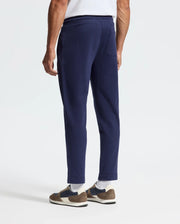 A person stands against a plain background, showcasing their lower half in Psycho Bunny's MENS ALEXANDER SWEATPANT - B6P155E200 in dark blue, with a relaxed fit and stylishly paired with brown and white sneakers.