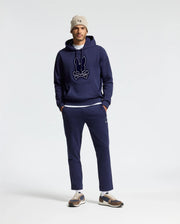 A man wearing a navy blue hoodie with a logo, Psycho Bunny's MENS ALEXANDER SWEATPANT - B6P155E200 in matching relaxed fit joggers, beige beanie, and sneakers stands against a plain background with his hands in his pockets.