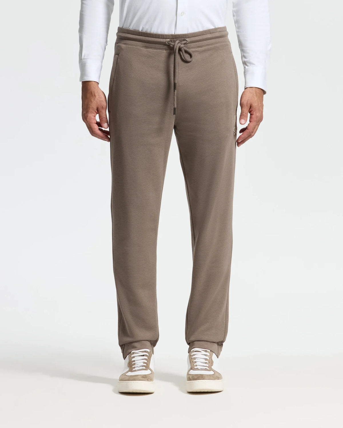A person stands against a neutral background wearing the MENS ALEXANDER SWEATPANT - B6P155E200 by Psycho Bunny in taupe, paired with white sneakers. These premium joggers boast a drawstring waist and side pockets, complemented by a white long-sleeve shirt for an effortless blend of comfort and style.