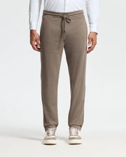A person stands against a neutral background wearing the MENS ALEXANDER SWEATPANT - B6P155E200 by Psycho Bunny in taupe, paired with white sneakers. These premium joggers boast a drawstring waist and side pockets, complemented by a white long-sleeve shirt for an effortless blend of comfort and style.