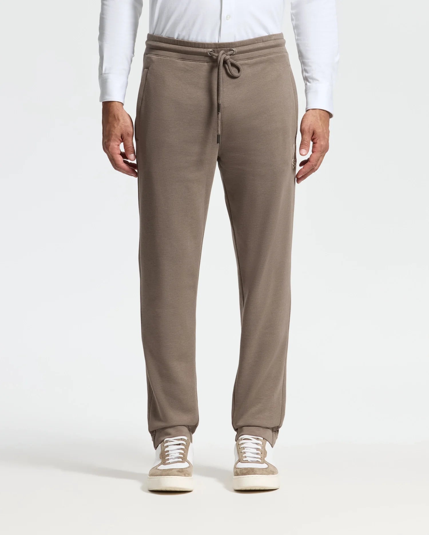 A person stands against a neutral background wearing the MENS ALEXANDER SWEATPANT - B6P155E200 by Psycho Bunny in taupe, paired with white sneakers. These premium joggers boast a drawstring waist and side pockets, complemented by a white long-sleeve shirt for an effortless blend of comfort and style.