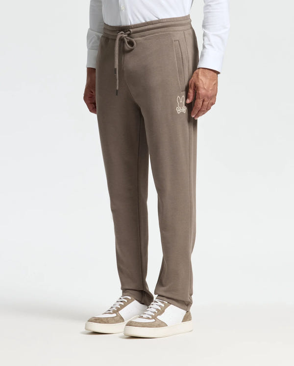 A person wearing the Psycho Bunny MENS ALEXANDER SWEATPANT - B6P155E200 in light brown, featuring a drawstring waistband and a small bunny logo on the left side, paired with white sneakers and a white shirt. The focus is on the premium quality joggers and footwear against a plain background.