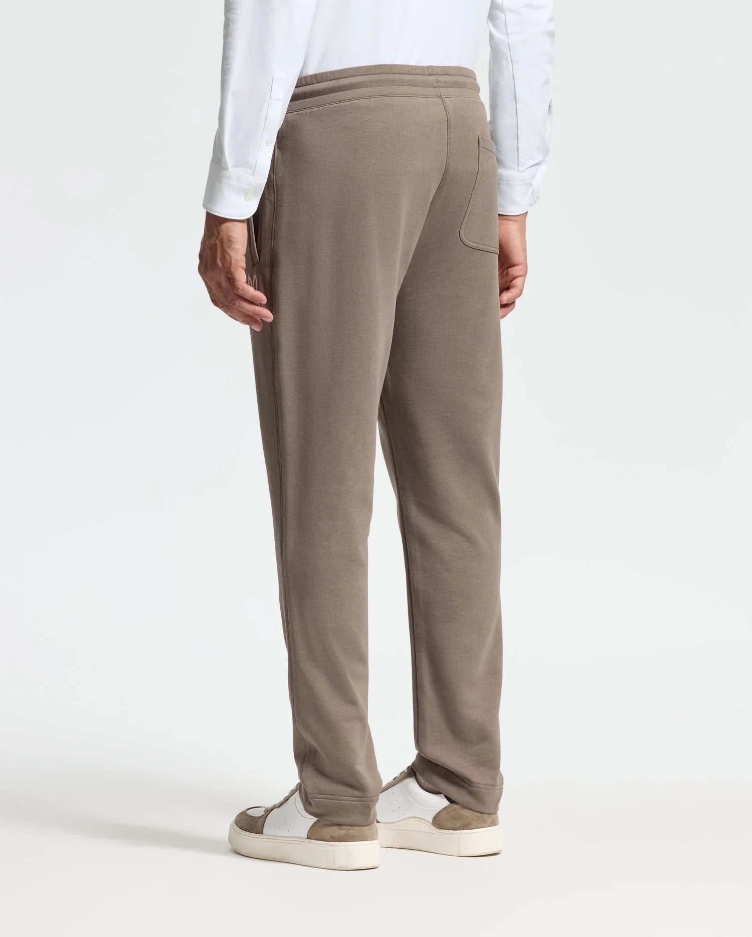 A person wearing a high-quality white long-sleeve shirt and Psycho Bunny's MENS ALEXANDER SWEATPANT - B6P155E200 in brown stands with their back to the camera. They pair this with white sneakers featuring beige soles, contributing to a relaxed look against the plain, light-colored background.