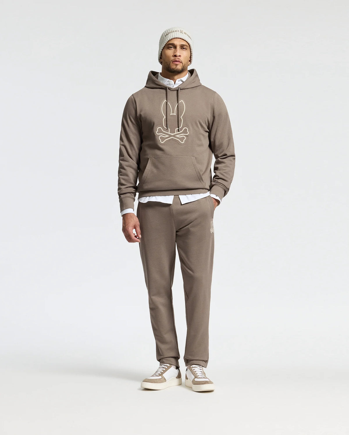A man stands against a plain background, wearing the premium quality taupe MENS ALEXANDER SWEATPANT - B6P155E200 by Psycho Bunny, along with a hoodie featuring a cartoon face design. He layers a white shirt underneath and complements the look with casual sneakers and a beanie.