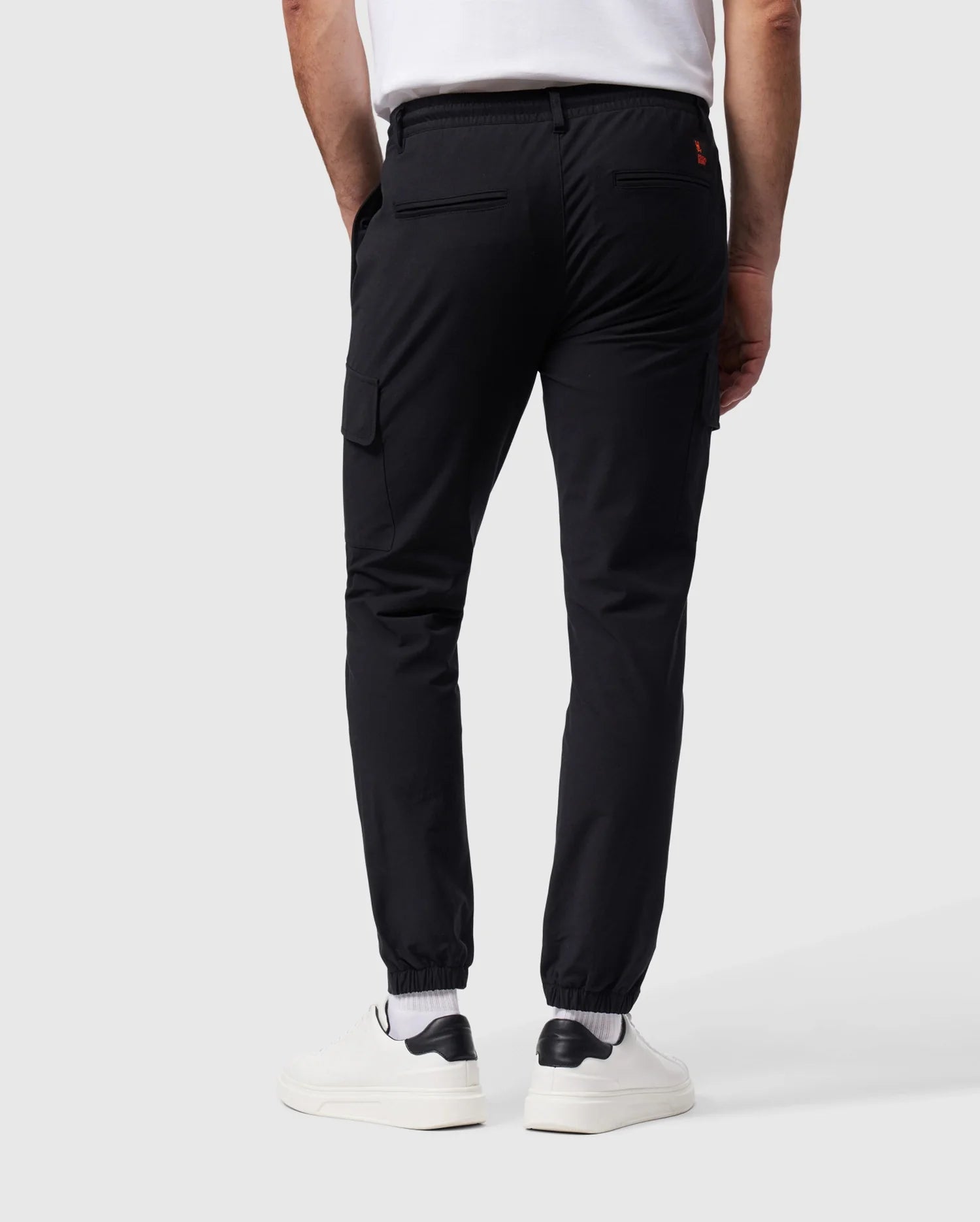 A person wearing Psycho Bunny's MENS LLOYD CARGO JOGGER - B6P159B200 in black, featuring elasticized cuffs and utilitarian pockets, paired with a white T-shirt, white socks, and white sneakers. The person is standing with their back facing the camera against a plain gray background.