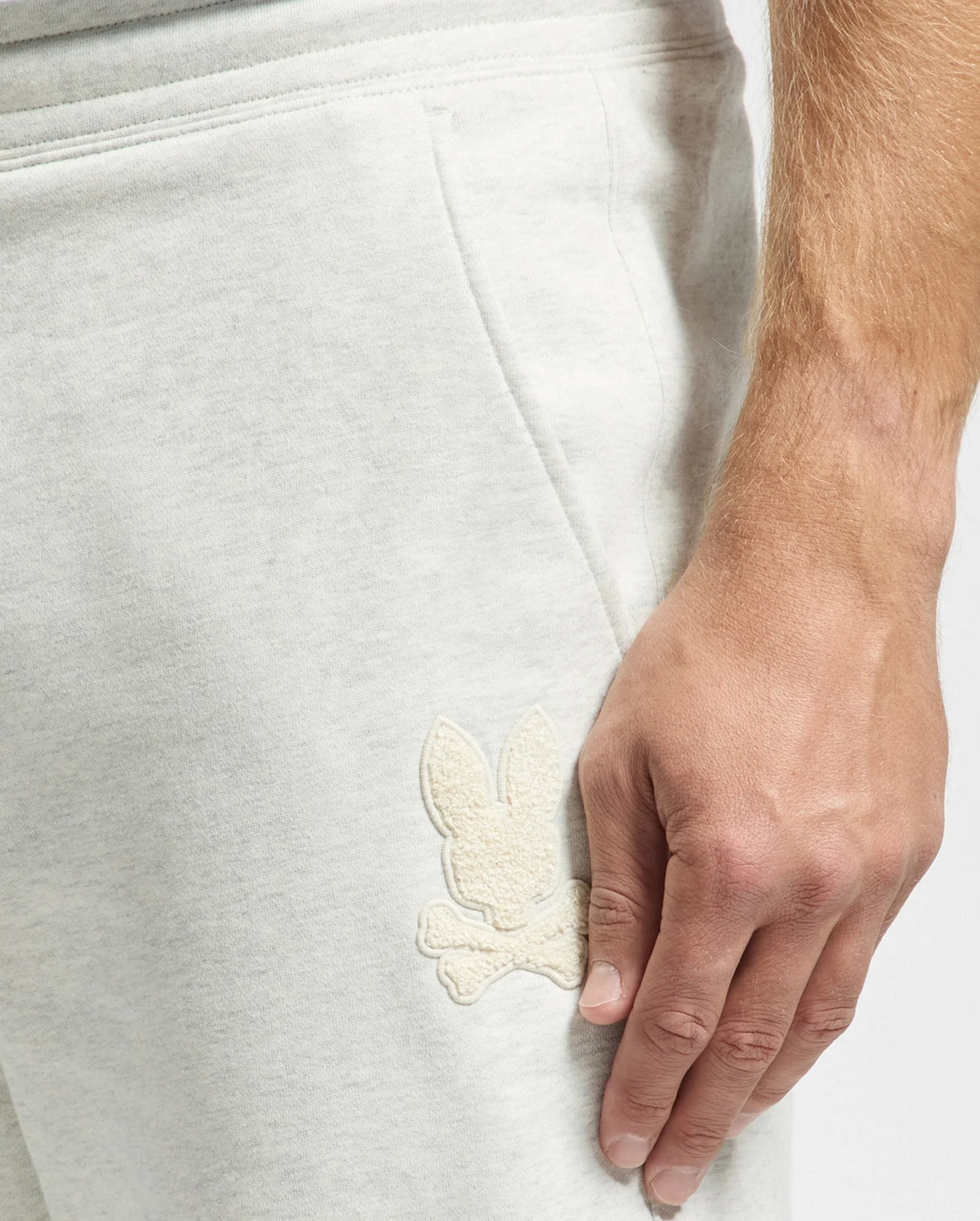 Close-up of a person wearing Psycho Bunny men's Eldorado joggers in light gray brushback cotton, featuring an embroidered Chenille Bunny logo above crossed bones on the pocket. A hand resting on the side highlights the detailed texture and emblem.