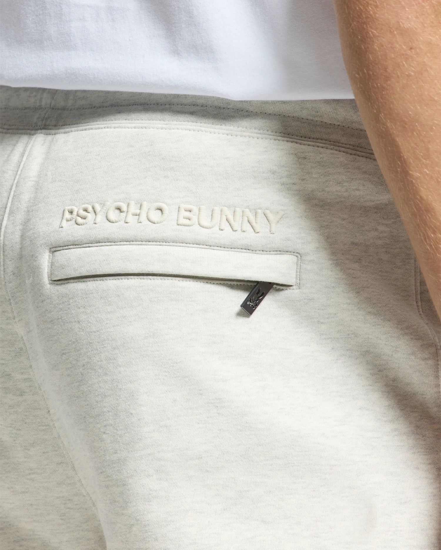 Close-up of the back pocket of light gray MENS ELDORADO BRUSH BACK JOGGER - B6P285E200 by Psycho Bunny with "Psycho Bunny" stitched above it. A small black tag near the seam features a Chenille Bunny logo. The side of a person in a white shirt is partially visible.