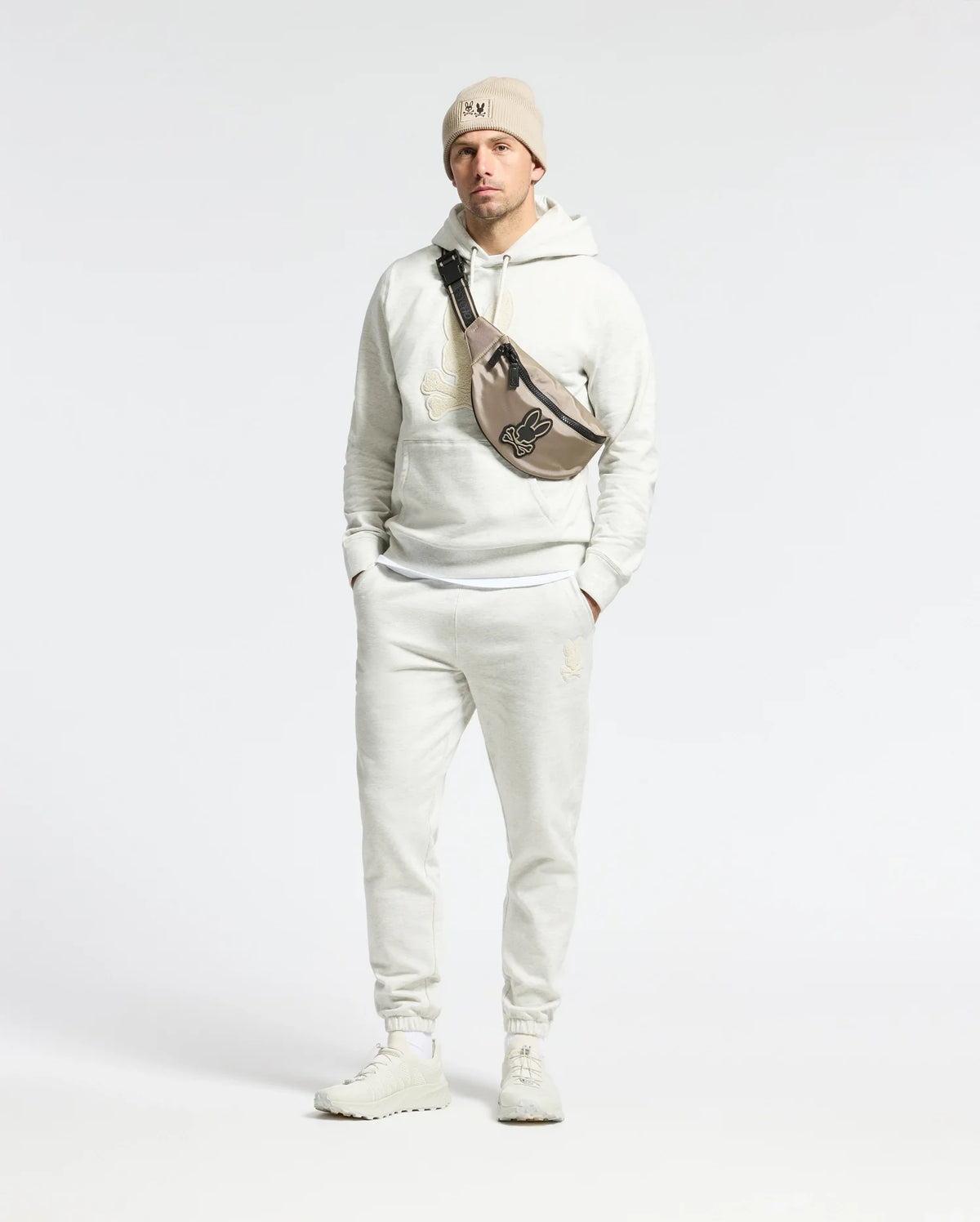 Sporting a Psycho Bunny Eldorado Brush Back Jogger, beanie, and sneakers, a person confidently stands with hands in pockets. Their crossbody bag features the Chenille Bunny logo against a plain backdrop.