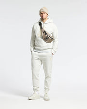 Sporting a Psycho Bunny Eldorado Brush Back Jogger, beanie, and sneakers, a person confidently stands with hands in pockets. Their crossbody bag features the Chenille Bunny logo against a plain backdrop.